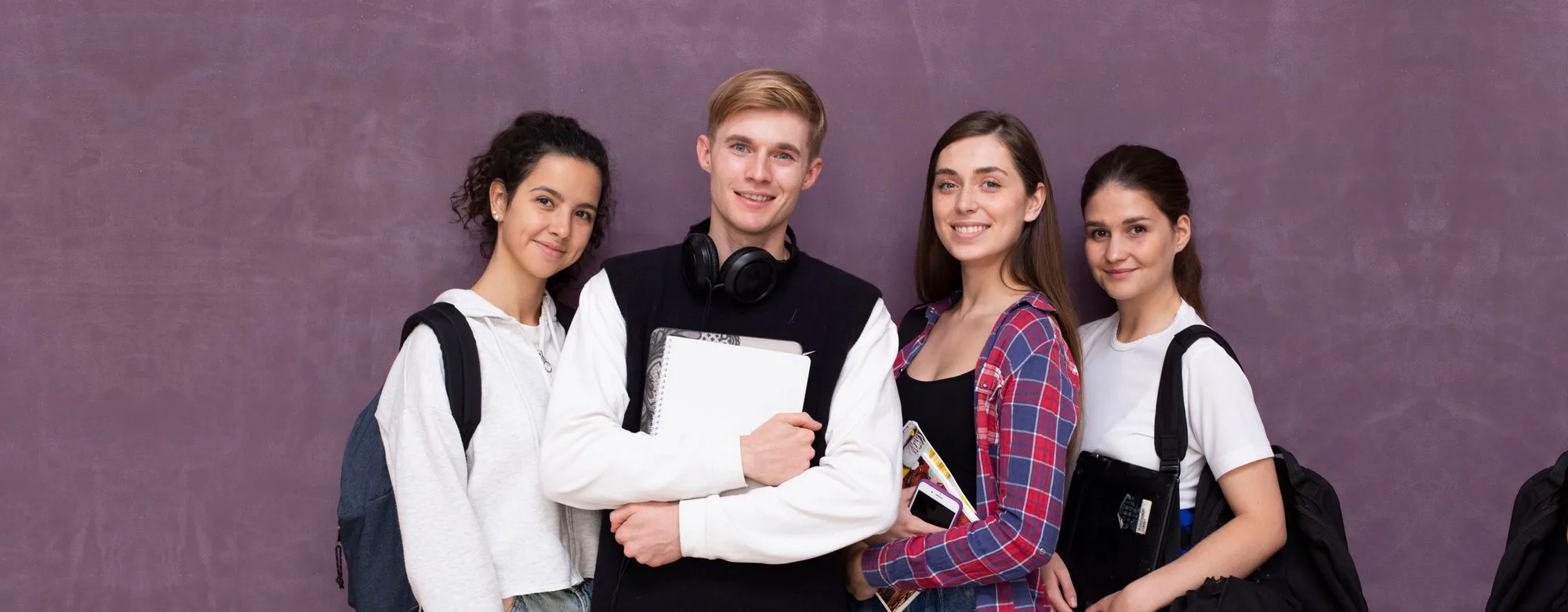 Four students with backpacks standing against a plain wall